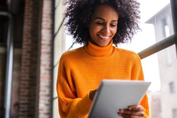 Woman in orange sweater in front of large window smiling as she looks at Shopify on her iPad