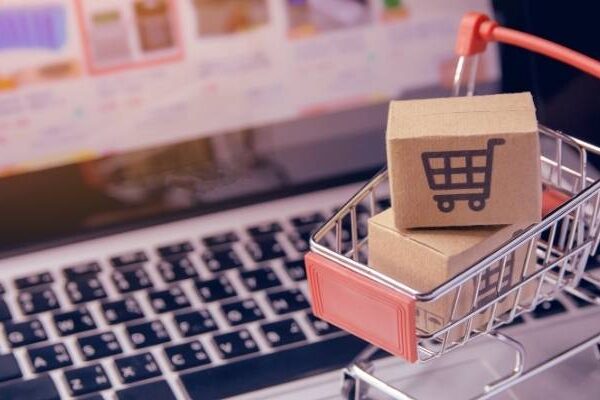 Tiny shopping cart filled with online purchases sitting in front of a laptop screen with ecommerce shopping in view