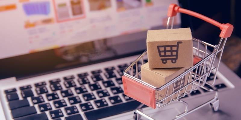Tiny shopping cart filled with online purchases sitting in front of a laptop screen with ecommerce shopping in view